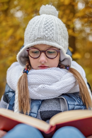 A warmly dressed teen girl reads a book outdoors.