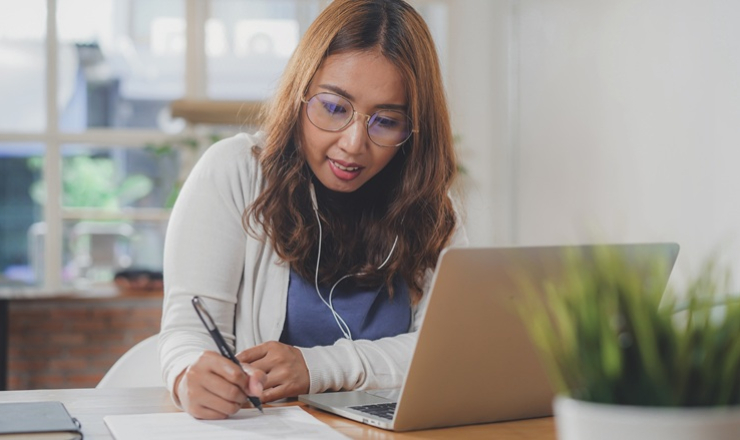 A pretty, Asian woman studies online with a laptop, earbuds, and a paper and pen.